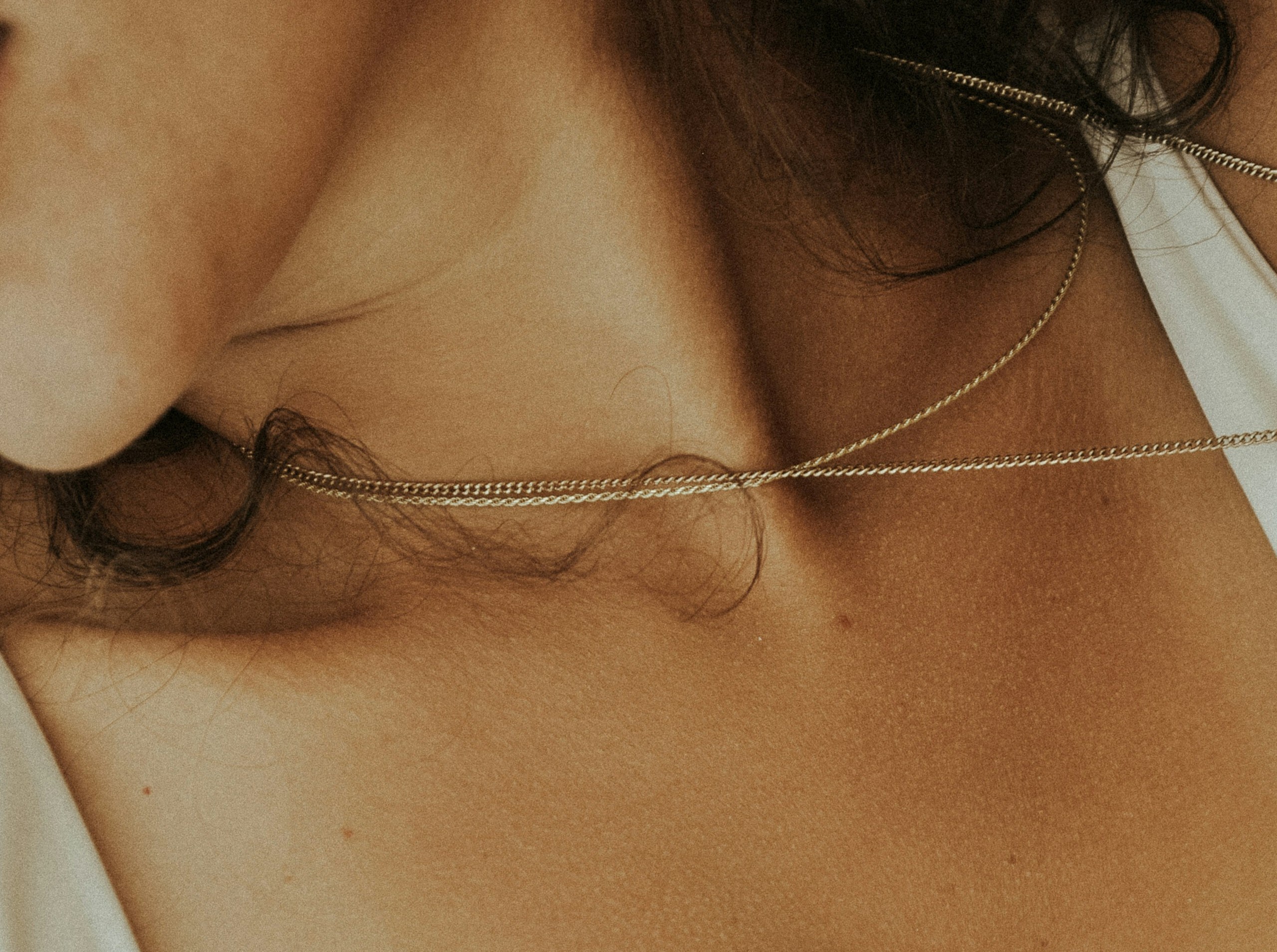 Close-up of a womans neck and collarbone, with two delicate gold chains resting on her skin and a few loose strands of hair visible. She is wearing a white top, evoking the relaxing ambiance of a facial spa.