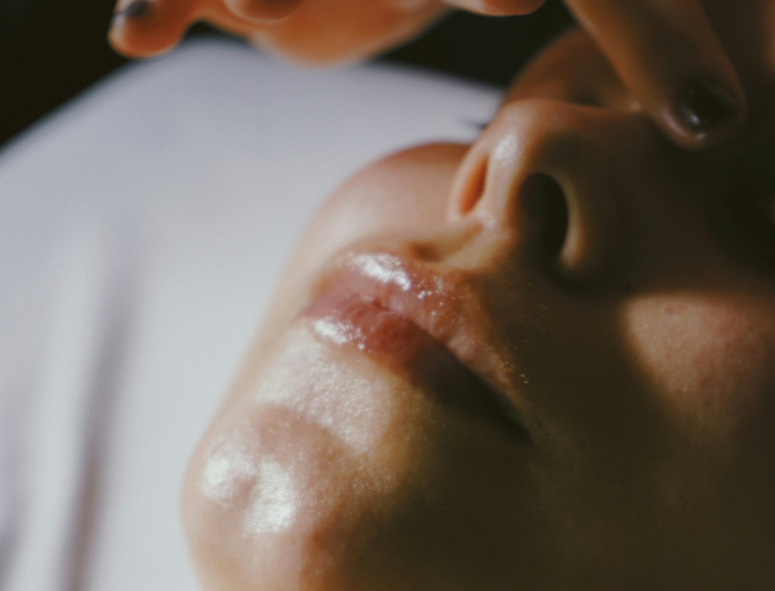 Close-up of a persons face lying down, with fingers gently touching the skin near the closed eye. The lighting is soft, highlighting the lips and chin. The mood appears calm and relaxed.