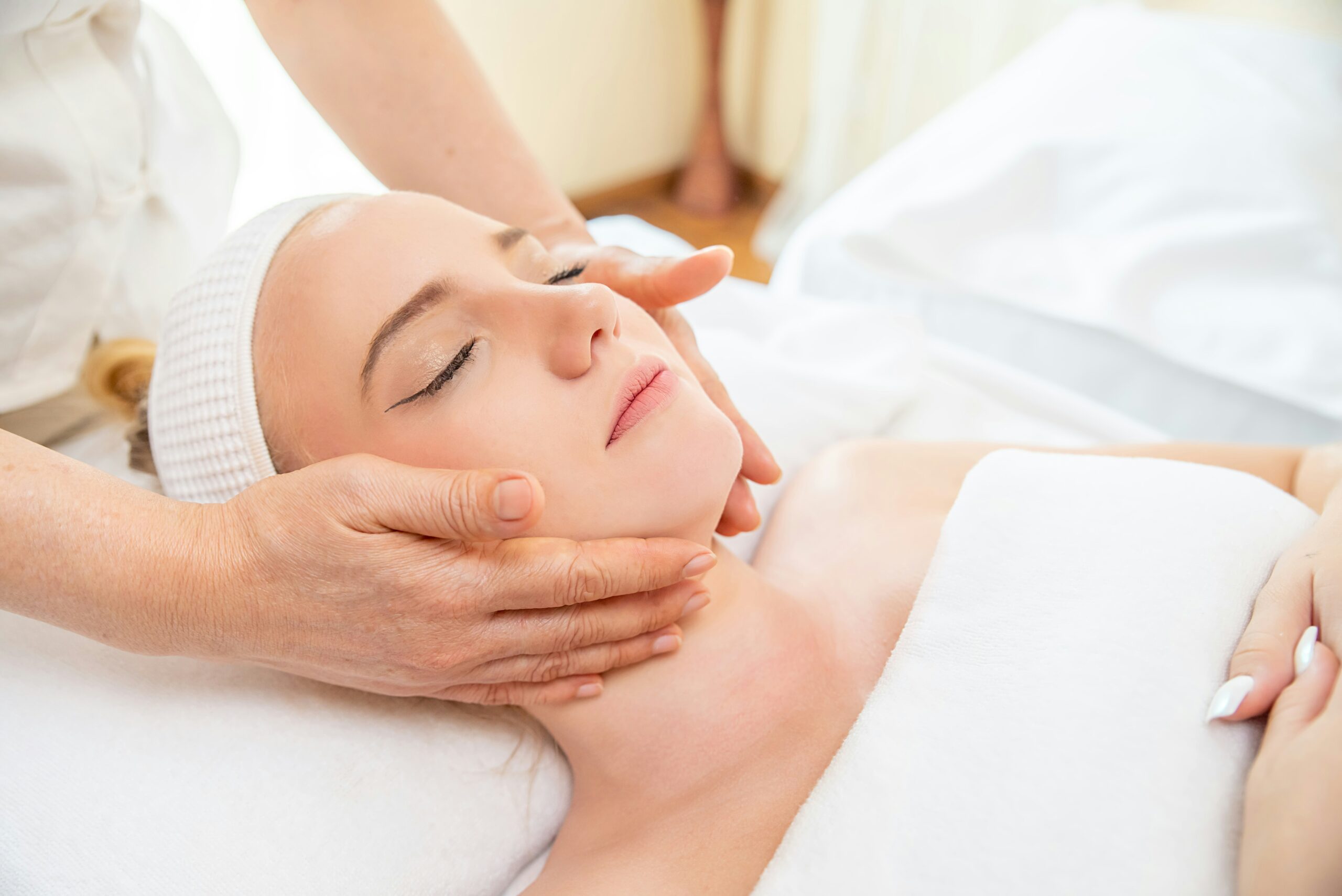 A person lying on a spa bed with eyes closed, covered in a white towel, receives a gentle facial treatment and massage from another person’s hands. The setting appears calm and relaxing.