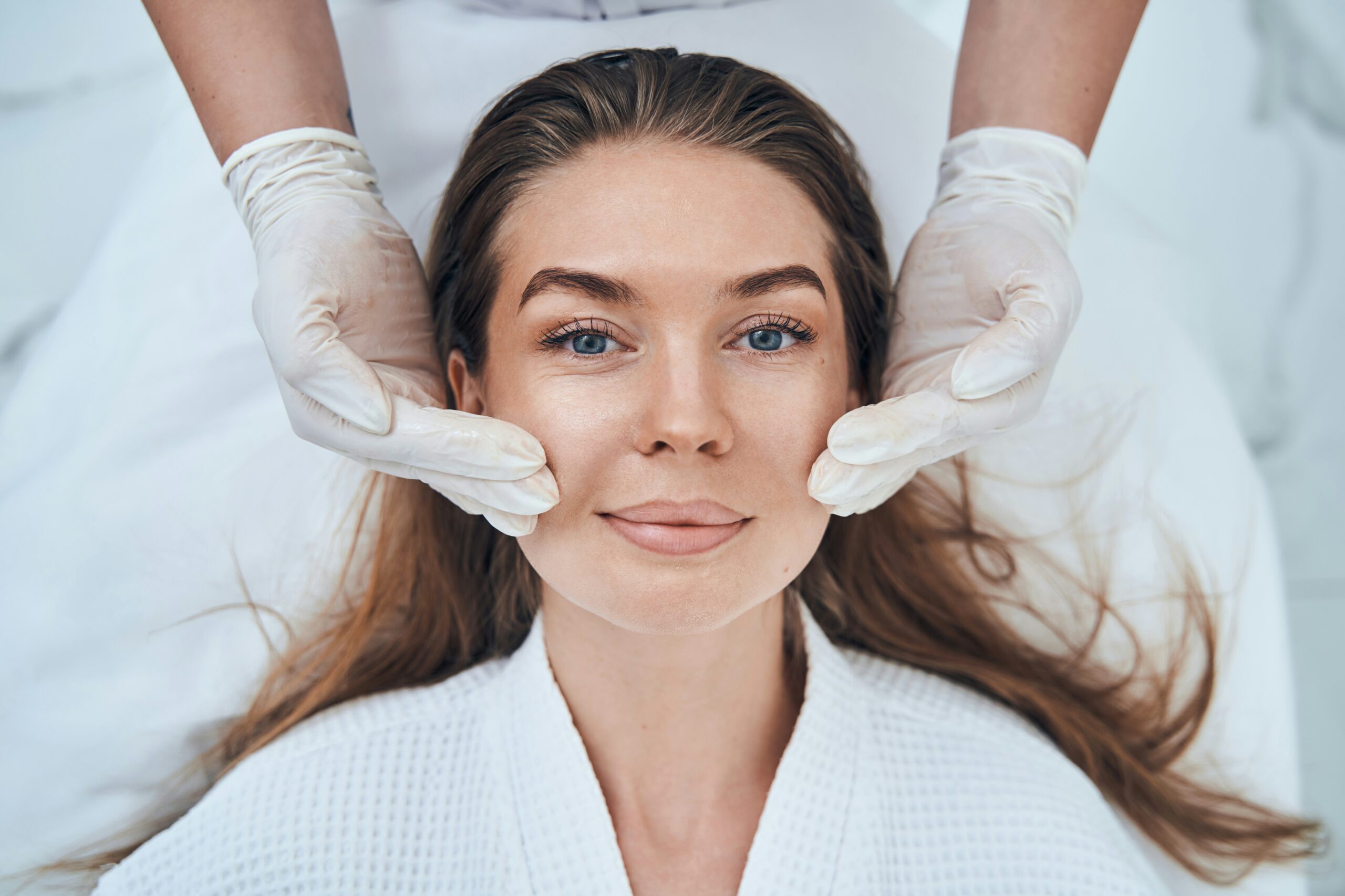 A woman with long hair lies on a spa table wearing a white robe. As part of her hormone therapy routine, a beautician in white gloves gently massages her face, with both hands on her cheeks. The woman looks relaxed and content.