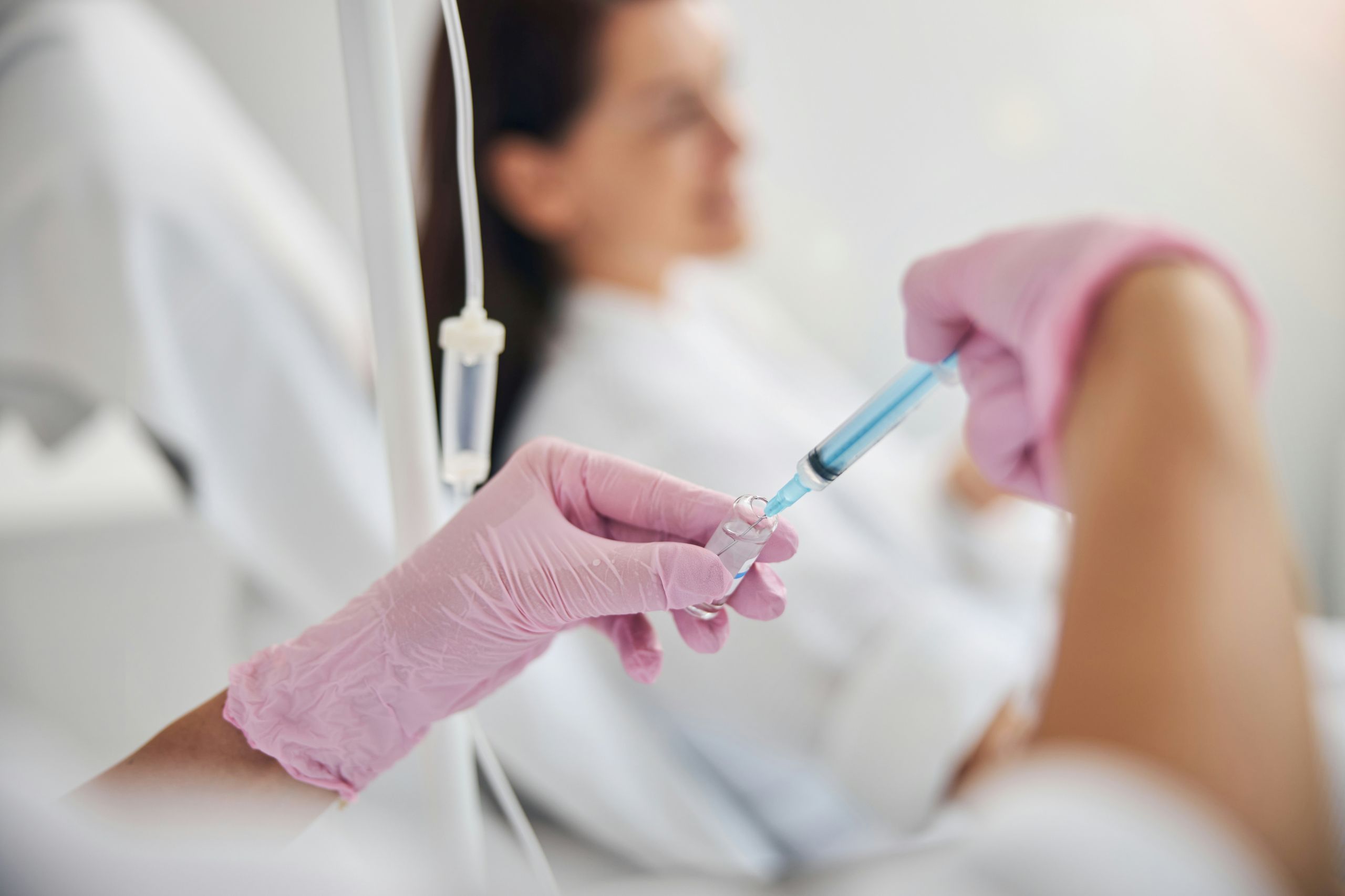 A healthcare worker wearing pink gloves prepares a syringe next to an IV bag, while a patient sits in the background, slightly out of focus.